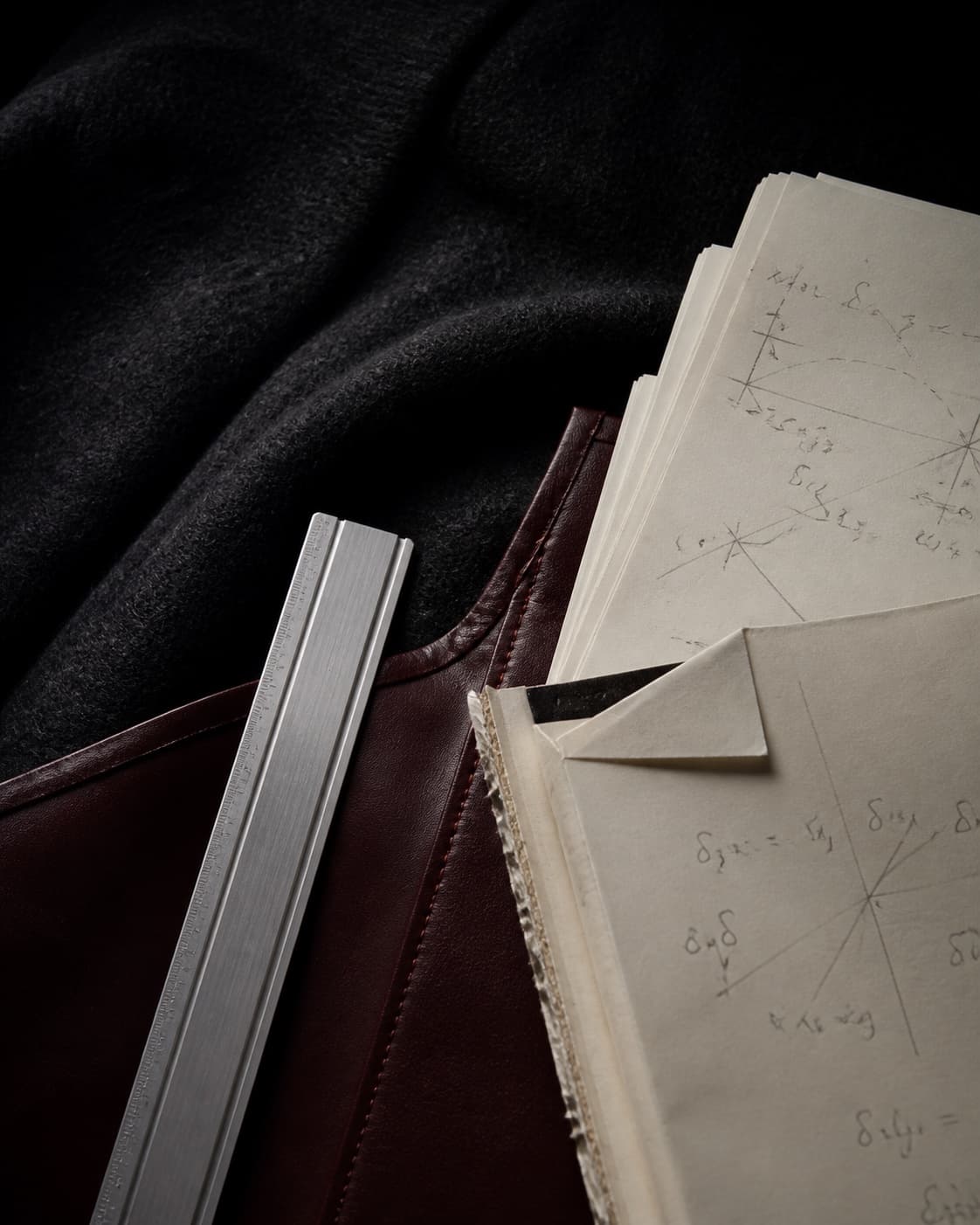 Ivory mathematics papers, an oxblood folio and a brushed aluminium ruler arranged on charcoal wool